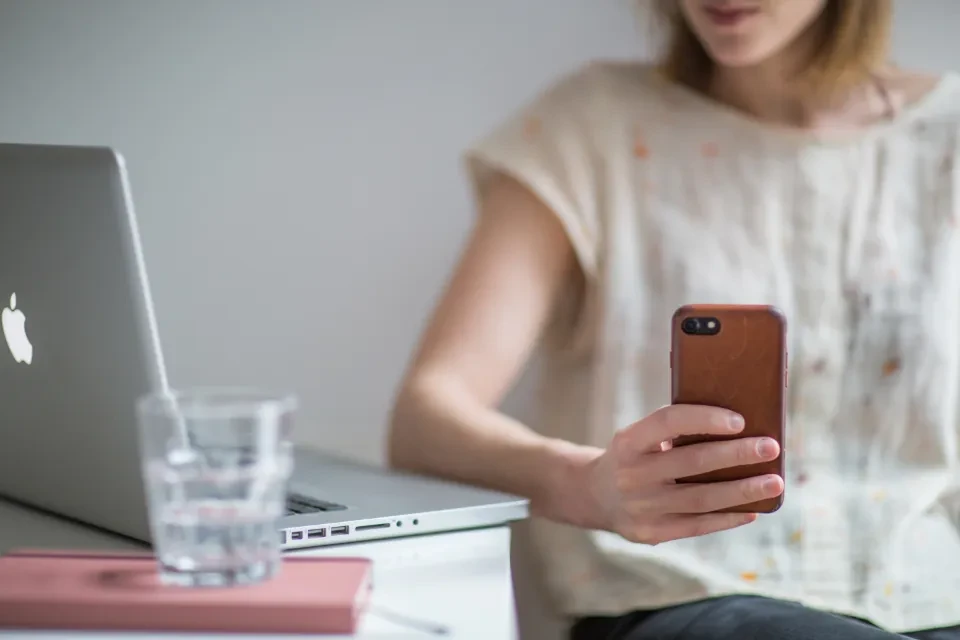 Una mujer consulta su teléfono móvil sentada a una mesa con un portátil y un vaso de agua