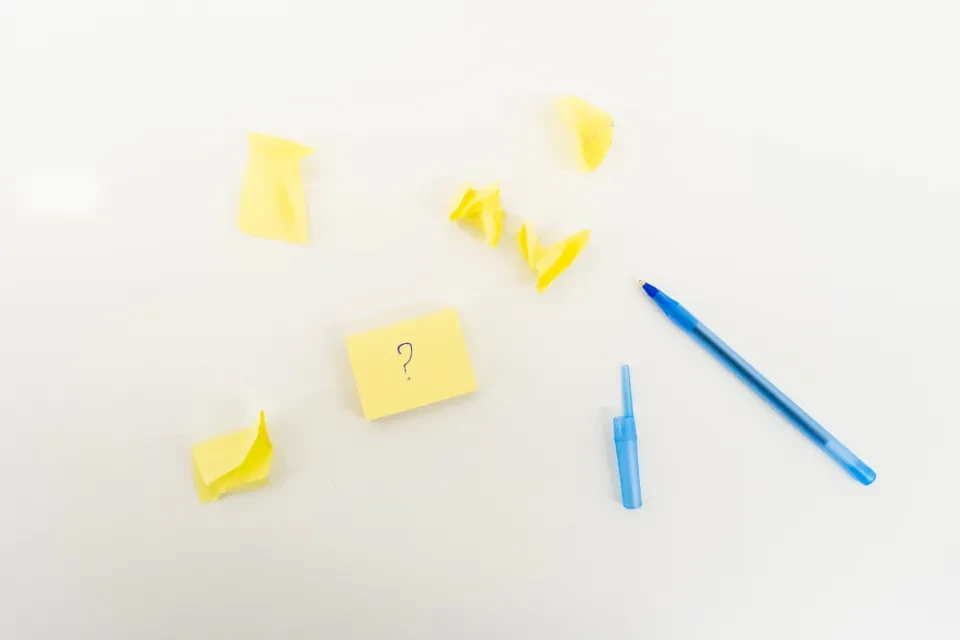 Notas adhesivas amarillas sobre una mesa blanca, una de ellas con un signo de interrogación, y dos bolígrafos azules