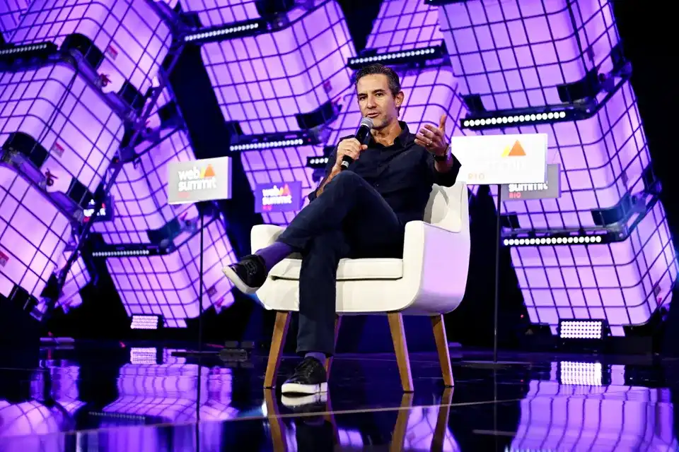 David Vélez, fundador de Nubank, habla con un micrófono sentado en un sillón blanco sobre un escenario iluminado en morado
