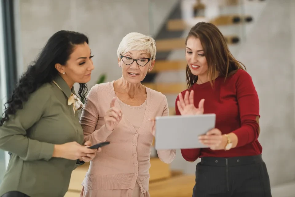Tres compañeras conversan de pie en una oficina; una de ellas enseña algo en una tableta a las otras dos