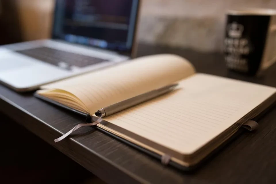 Un cuaderno abierto con las páginas en blanco junto a un portátil sobre una mesa de madera oscura
