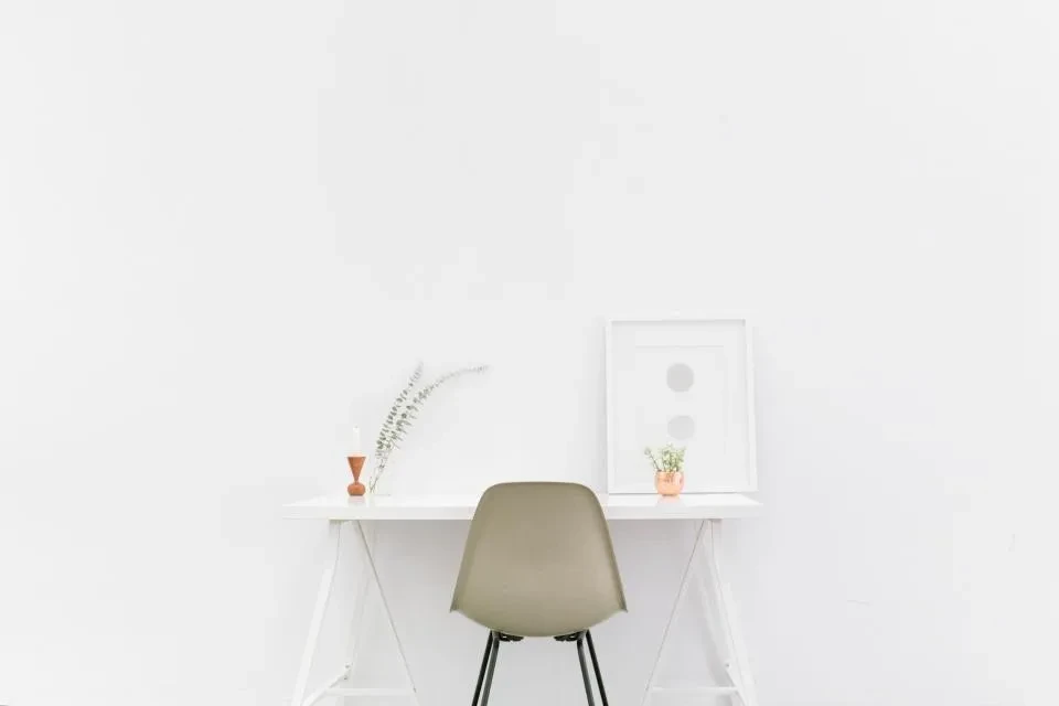 Un escritorio blanco con una silla y una planta, preparado y vacío junto a una pared blanca